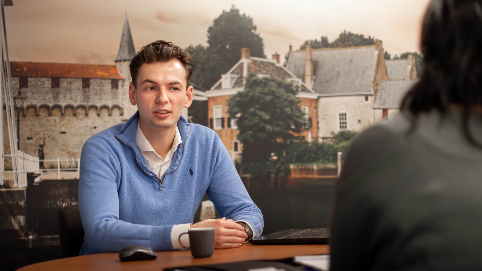Business consultant van Invision ICT in gesprek met een klant aan tafel, met een fotowand van een historische stadspoort aan het water op de achtergrond